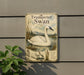 A metal sign with a picture of a swan and text about the trumpet swan, mounted on a wall next to a plant.
