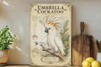 a vintage-style illustration of an umbrella cockatoo, a large white bird with a distinctive crest, perched on a branch. The illustration is accompanied by text that provides information about the bird, including its species, habitat, and native range. The image is displayed on a wooden shelf, with a potted plant and a wooden cutting board visible in the background.