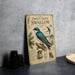 a vintage-style metal sign with illustrations and text about a violet-green swallow, placed on a table next to some black camera lenses.
