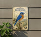 a decorative sign depicting a bluebird perched on a tree stump, surrounded by pine cones and other natural elements.
