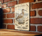 A vintage-style metal plaque featuring an illustration of a bird, likely a Whimbrel, standing on a rocky shoreline with a brick wall in the background.