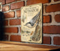 a vintage-style illustration of a white-breasted nuthatch perched on a tree branch, mounted on a brick wall.
