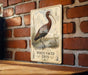A vintage-style metal sign featuring an illustration of a white-faced ibis bird standing on a brick wall.