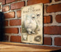 a vintage-style illustration of a white Silkie chicken, which is a breed of chicken known for its distinctive appearance and elegant demeanor. The illustration is mounted on a brick wall, and there is a wooden table below it. The image does not contain any text, and it does not depict any actions or movements.