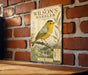 A vintage-style metal plaque featuring a yellow warbler bird on a branch, with a map and text below it, mounted on a brick wall.