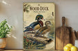 A framed illustration of two wood ducks perched on a log, surrounded by a natural scene, is displayed on a shelf next to a wooden cutting board and some fruit.