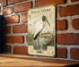 A vintage-style metal sign featuring an illustration of a wood stork standing in a marshy area, mounted on a brick wall.