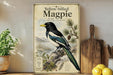 A vintage-style poster of a yellow-billed magpie is displayed on a shelf, accompanied by a potted plant, a wooden cutting board, and some lemons.