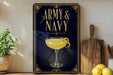 A framed poster with the text "ARMY & NAVY" and an illustration of a cocktail glass with a lemon twist, placed on a wooden shelf next to a cutting board and a potted plant.