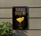 A sign for a banana daiquiri cocktail is displayed on a wall, with a plant visible to the left.