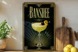 A framed poster of a cocktail with a lemon twist and a cherry on top, placed on a wooden shelf next to a cutting board and a potted plant.