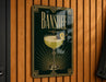 A decorative sign for "Banshee" cocktail, featuring a glass of yellow liquid with a lemon twist and a cherry on top, mounted on a wooden wall.