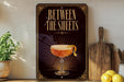 A decorative sign with the text "BETWEEN THE SHEETS" and an illustration of a cocktail glass with a lemon twist.