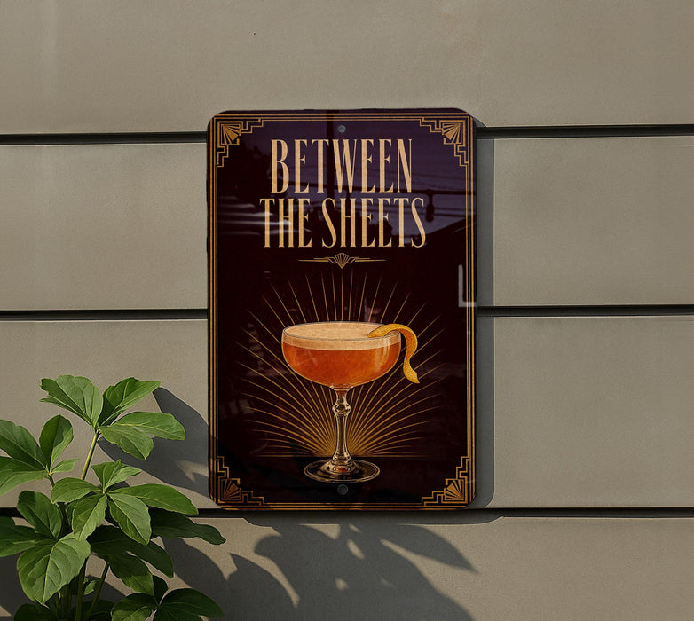 A sign with the text "BETWEEN THE SHEETS" and an illustration of a cocktail glass with a lemon peel on top, mounted on a wall next to a plant.