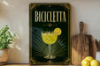 a decorative sign with the word "BICICLEETTA" on it, along with an illustration of a glass containing a yellow drink and a lemon slice. The sign is displayed on a shelf, surrounded by various kitchen items such as a wooden cutting board, a lemon, and a potted plant.