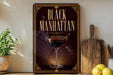 a decorative sign with the text "Black Manhattan" and an illustration of a martini glass with a cherry on top, placed on a wooden surface next to a cutting board and a potted plant.