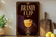 A dark wooden sign with a glass illustration of a cocktail, accompanied by the text "BRANDY FLIP" in a decorative font. The sign is displayed on a shelf, with a potted plant, a wooden cutting board, and some lemons nearby.