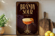 A decorative sign for a Brandy Sour cocktail, featuring an illustration of a glass with a lemon twist, placed on a wooden surface next to a cutting board and some lemons.