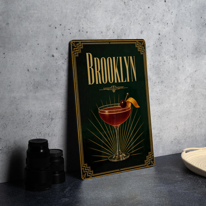 A framed poster of a cocktail with the name "Brooklyn" on it, placed on a gray countertop next to some black camera lenses.