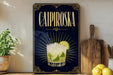 a decorative sign with the text "CAIPIROSKA" and an illustration of a cocktail glass with a lime slice. The sign is displayed on a wooden shelf, with a potted plant, a cutting board, and some lemons nearby.