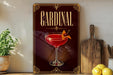 A decorative sign for a "Cardinal" cocktail, featuring a glass of red cocktail with a cherry on top, is displayed on a wooden shelf next to a cutting board and some lemons.