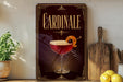 A decorative sign for "Cardinale" cocktail, featuring a glass of red cocktail with an orange twist and garnish, placed on a wooden surface next to a cutting board and a potted plant.
