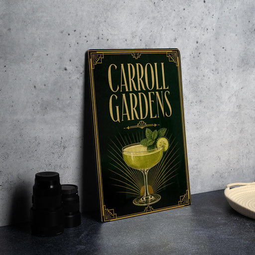 A decorative sign for "Carroll Gardens" featuring a cocktail glass and mint leaves, placed on a gray countertop next to some black camera lenses.