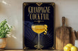 A framed advertisement for a champagne cocktail, featuring a glass of champagne with a lemon twist, is displayed on a wall next to a wooden cutting board and a potted plant.