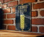 A sign for a cucumber Collins cocktail is displayed on a brick wall.