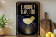 A decorative sign for "Florida Daiquiri" featuring a cocktail glass with a lime slice, placed on a wooden surface next to a wooden cutting board and a potted plant.