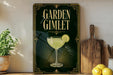 A framed advertisement for a "Garden Gimlet" cocktail, featuring a glass of the drink with a slice of lemon, is displayed on a shelf next to a wooden cutting board and a potted plant.
