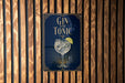 A sign with the text "GIN AND TONIC" and an illustration of a gin and tonic glass, placed on a wooden wall.