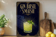 A framed poster advertising a Gin Basil Smash cocktail, featuring a glass of the drink and a basil leaf, is displayed on a wall next to a wooden cutting board and a potted plant.