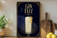 A decorative sign with the text "GIN Fizz" and an illustration of a cocktail glass with a lemon twist, placed on a wooden surface next to a potted plant and a cutting board.