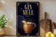 A framed poster with the text "GIN MULE" and an illustration of a copper mug with a lemon slice, placed on a wooden shelf next to a wooden cutting board and a potted plant.