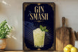 A decorative sign with the text "GIN SMASH" and an illustration of a cocktail, placed on a wooden surface next to a wooden cutting board and a potted plant.