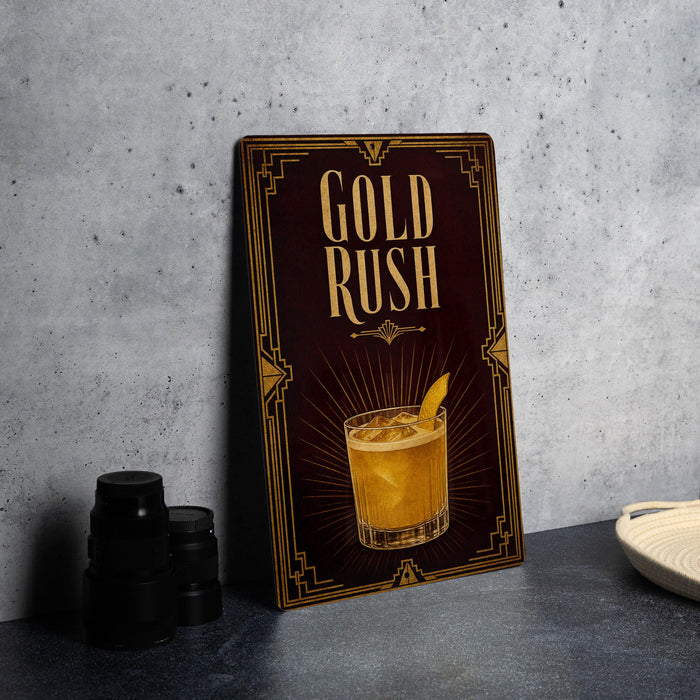 A decorative sign with the text "GOLD RUSH" and an illustration of a cocktail, placed on a gray countertop next to some black containers.