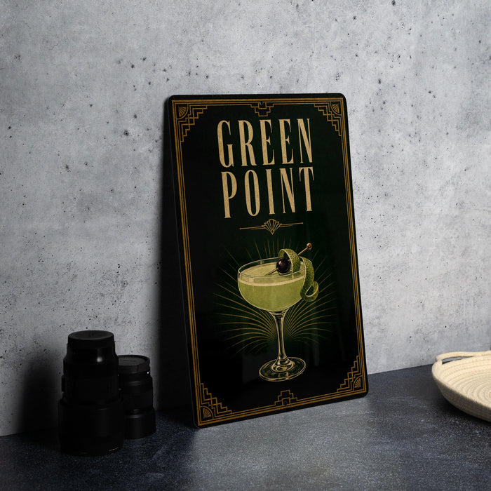 A framed poster of a cocktail with a green drink and a cherry on top, placed on a gray countertop next to a stack of black camera lenses.