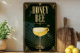 A framed poster with the text "HONEY BEE" and an illustration of a cocktail glass with a lemon twist, placed on a wooden shelf next to a wooden cutting board and a potted plant.