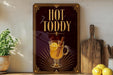 A framed advertisement for "Hot Toddy" featuring a glass of hot toddy with a lemon slice and cinnamon stick, placed on a wooden shelf next to a cutting board and a potted plant.