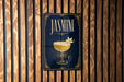 A sign with the word "JASMINE" and an illustration of a cocktail glass with a flower on it, mounted on a wooden wall.