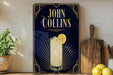 A poster advertising a drink called "John Collins" with a glass of lemonade and a lemon slice, placed on a wooden surface next to a wooden cutting board and a potted plant.