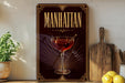 A framed poster of a Manhattan cocktail is displayed on a wall, with a wooden cutting board and a potted plant to the side.