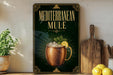 a poster or sign with the text "MEDITERRANEAN MULE" and an illustration of a copper mug with a lemon slice and mint leaves. The poster is displayed on a wall, and there are some kitchen items, such as a wooden cutting board and a potted plant, in the foreground.