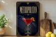 a poster or sign with the word "METROPOLITAN" prominently displayed, along with an illustration of a martini glass containing a red drink and a lemon twist. The poster is set against a dark background with a decorative border, and there are some potted plants and kitchen utensils visible in the foreground.