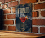 A framed poster of a cocktail, featuring a red drink in a martini glass, is displayed on a brick wall.