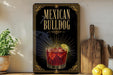 A decorative sign for "Mexican Bulldog" cocktail, featuring a glass of red liquid with ice and a lime wedge, is displayed on a wooden shelf next to a wooden cutting board and a potted plant.