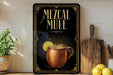A framed advertisement for a mezcal mule, featuring an illustration of a copper mug with a lime slice, placed on a wooden shelf next to a wooden cutting board and a potted plant.