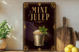 A framed poster with the text "MINT JULEP" and an illustration of a mint julep drink, surrounded by decorative elements and a wooden cutting board.