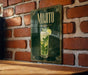 A framed poster of a mojito cocktail is displayed on a brick wall, with a wooden table below it.